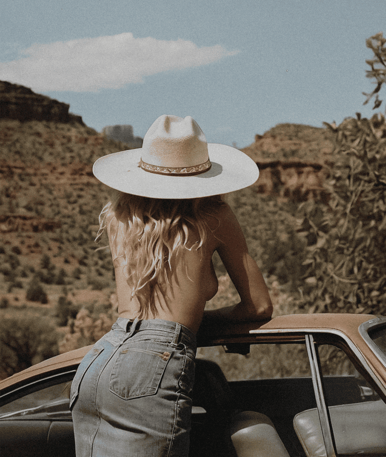 Person wearing a wide-brimmed hat and high-waisted jeans standing by a car in a desert landscape.